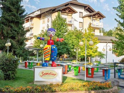 Familienhotel - Kinderbecken - Kärnten - Maskottchen Sonnelino mit Hotel und Pit Pat im Hintergrund - Baby + Kinderhotel Sonnelino