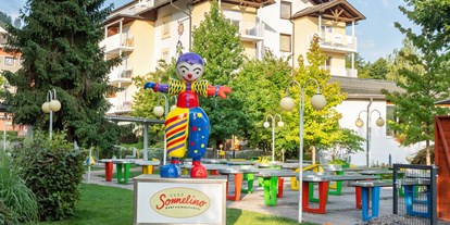 Familienhotel - Kinderbetreuung - Keutschach - Maskottchen Sonnelino mit Hotel und Pit Pat im Hintergrund - Baby + Kinderhotel Sonnelino