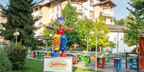 Familienhotel - Kinderhotels Europa - Österreich - Maskottchen Sonnelino mit Hotel und Pit Pat im Hintergrund - Baby + Kinderhotel Sonnelino