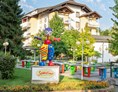 Kinderhotel: Maskottchen Sonnelino mit Hotel und Pit Pat im Hintergrund - Baby + Kinderhotel Sonnelino